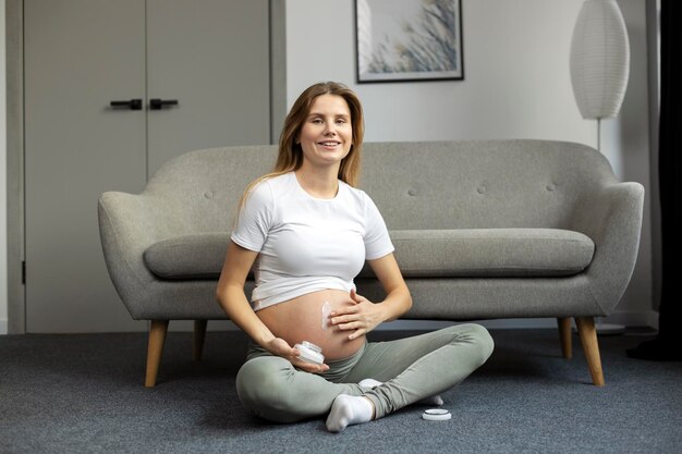 Lächelnde schwangere Frau, die zu Hause sitzt und Creme auf den Bauch aufträgt. Schwangerschaft, Hautpflegekonzept
