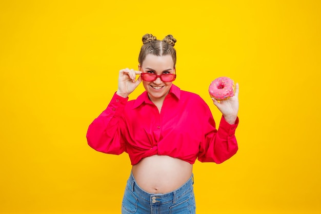 Lächelnde schwangere Frau in rosa Hemd mit köstlichen süßen Donuts auf isoliertem gelbem Hintergrund. Süßes, kalorienreiches Essen während der Schwangerschaft. Eine schwangere Frau isst fetthaltiges Gebäck