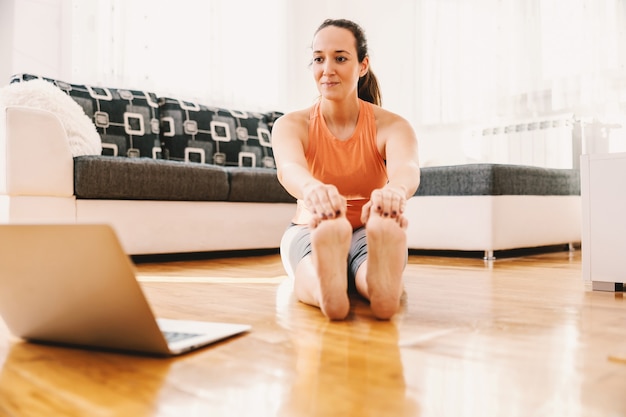 Lächelnde schwangere Sportlerin, die zu Hause auf dem Boden sitzt, Dehnübungen macht und einem Online-Kurs folgt