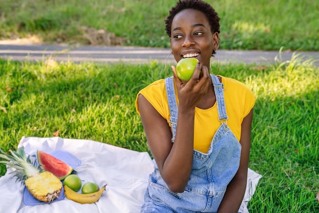 lächelnde schwarzafrikanische Frau, die sitzt und einen Apfel in einem Park im Freien isst, während sie ein Obstpicknick hat