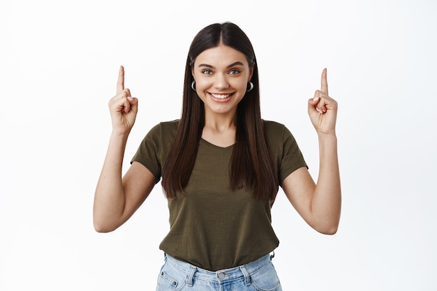 Lächelnde selbstbewusste Frau, die Werbung oben zeigt, mit den Fingern nach oben zeigt und fröhlich nach vorne schaut, einen Hinweis gibt, weiße Wand