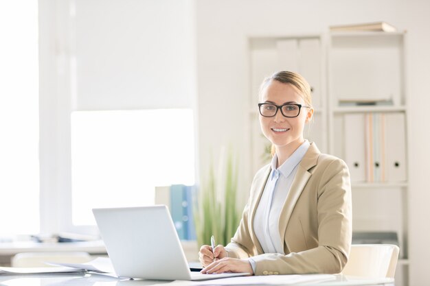 Lächelnde selbstbewusste hübsche junge Geschäftsdame in Brillen, die am Tisch mit Laptop sitzen und Notizen machen, während sie im Büro arbeiten