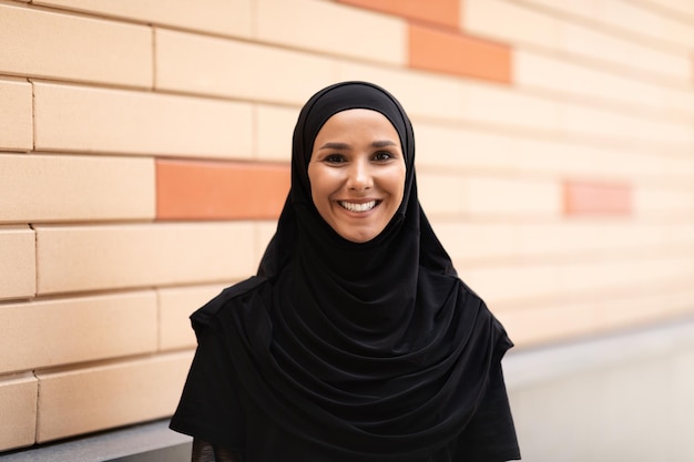 Lächelnde, selbstbewusste, hübsche junge islamische Frau im Hijab, die auf Backsteinmauerhintergrund in die Kamera blickt