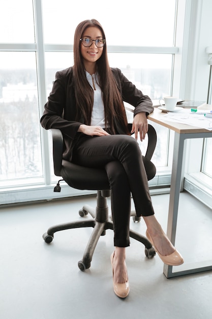 Lächelnde selbstbewusste junge Geschäftsfrau, die am Tisch im Büro sitzt