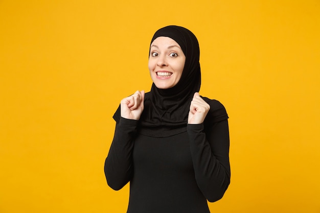Lächelnde selbstbewusste schöne junge arabische muslimische Frau in Hijab schwarzer Kleidung posiert isoliert auf gelber Wand, Porträt. Menschen religiösen Islam Lifestyle-Konzept.