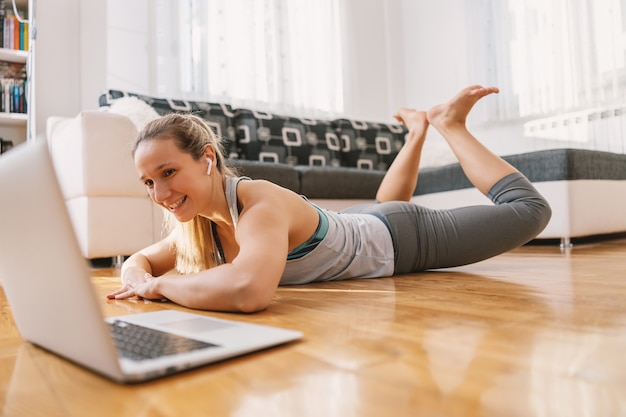 Lächelnde Sportlerin, die auf dem Bauch liegt und Rückenübungen macht, während sie dem Online-Unterricht während der Quarantäne folgt.