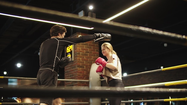 Foto lächelnde sportlerin macht schläge auf den boxring. männlicher boxer gibt anweisungen im fitnessstudio. schönes, fittes mädchen trainiert mit trainer im sportverein