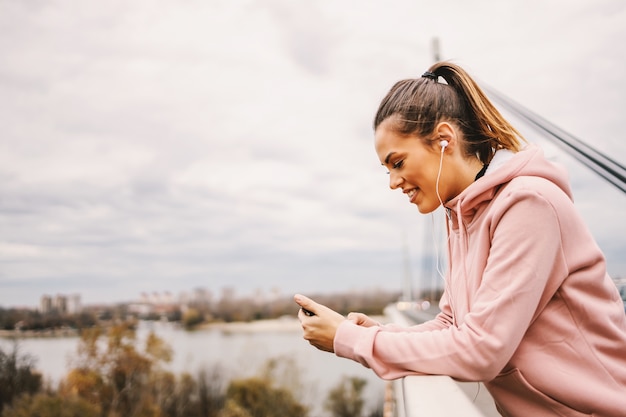 Lächelnde Sportlerin mit gesunden Gewohnheiten, die eine Pause nach dem Laufen und SMS auf dem Smartphone machen. Städtisches Äußeres.