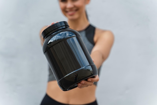 Foto lächelnde sportliche frau, die ein glas protein an ihren händen über grauem hintergrund hält und in die kamera lächelt fokus auf das schwarze glas