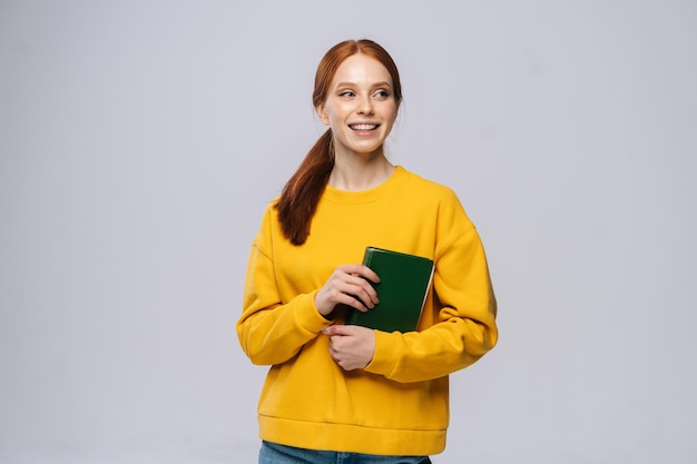 Lächelnde Studentin der jungen Frau, die gelben Pullover trägt, Buch hält und verträumt wegschaut