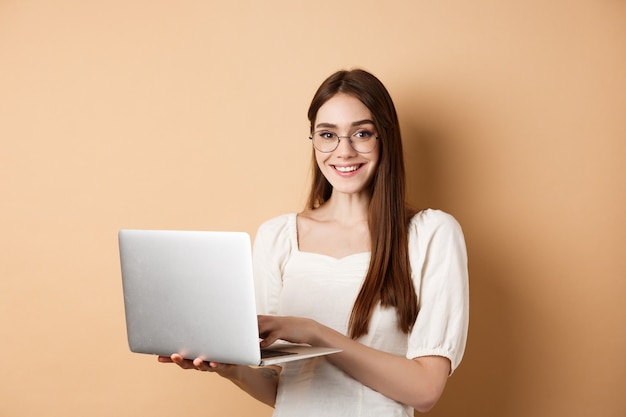 Lächelnde Studentin, die am Laptop arbeitet, eine Brille trägt und glücklich aussieht, Computer benutzt, während sie gegen Beige steht.