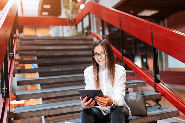 Lächelnde Studentin mit Brille und braunem Haar unter Verwendung der Tablette beim Sitzen auf der Treppe.