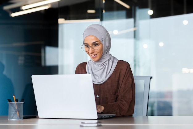 Lächelnde und verträumte Geschäftsfrau, die im Büro mit Laptop-Frau im Hijab und Brillenbüro arbeitet