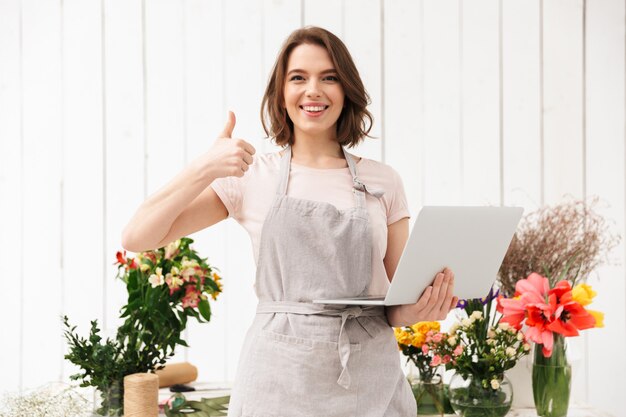 Lächelnde Verkäuferin, die nahe Blumensträußen im Blumenladen steht und Daumen oben mit Laptop in der Hand zeigt