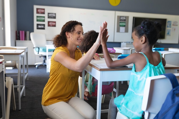 Foto lächelnde weiße junge lehrerin, die einem afroamerikanischen mädchen, das am schreibtisch sitzt, highfive gibt