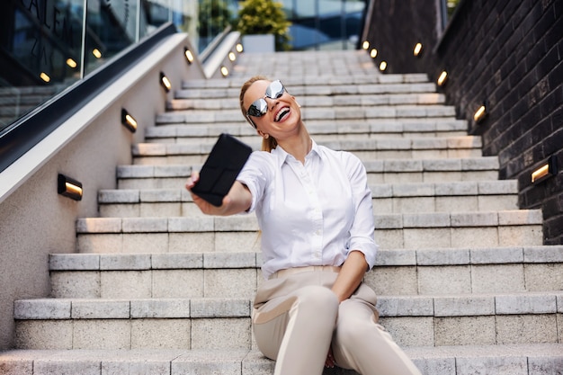 Lächelnde wunderschöne blonde modische Geschäftsfrau, die auf der Treppe vor Geschäftszentrum sitzt und selfie nimmt.