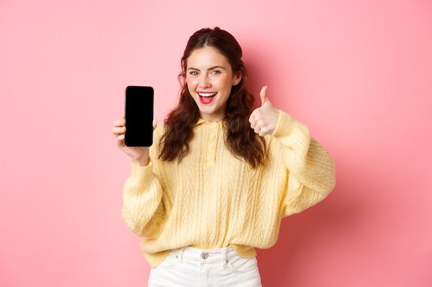 Lächelnde zufriedene Frau, die Daumen hoch und leeren Smartphone-Bildschirm zeigt, App oder Website empfiehlt, an rosa Wand stehend.