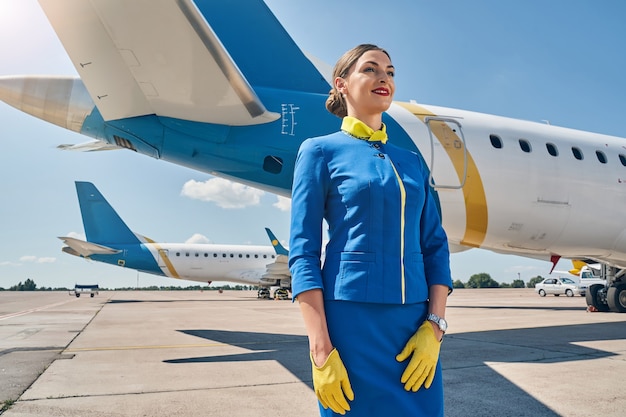Lächelnde zufriedene junge Frau in einer trendigen Airline-Uniform und Lederhandschuhen, die in die Ferne blicken