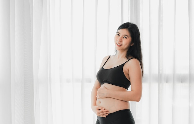 Lächelnde zwei Monate schwangere Asiatin berührt ihren Bauch am Fenster
