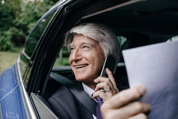 Lächelnder älterer Geschäftsmann mit Dokumenten am Telefon in einem Auto