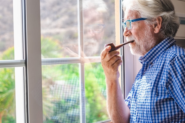 Lächelnder älterer Mann am Fenster mit Blick nach draußen beim Pfeifenrauchen