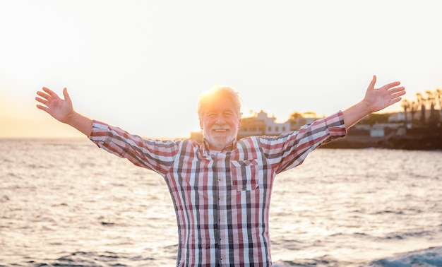 Lächelnder älterer Mann am Meeresstrand bei Sonnenuntergang, der mit ausgestreckten Armen in die Kamera blickt