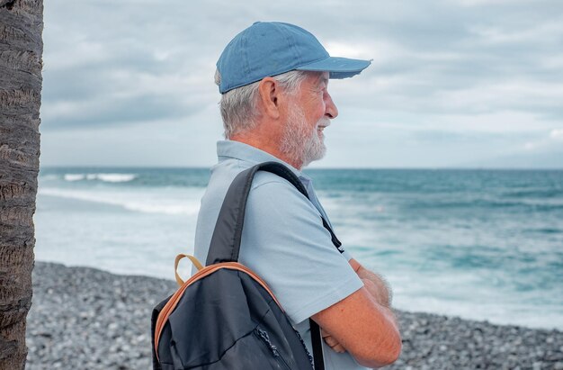 Lächelnder älterer Mann, der am Strand in der Nähe einer Palme steht und Freizeiturlaub oder Ruhestand genießt Attraktiver bärtiger Mann mit Hut und weißem Haar, der den Horizont betrachtet