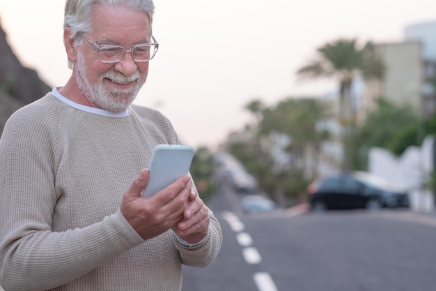 Lächelnder älterer Mann, der auf der Straße steht und auf das Handy schaut und Technik und Soziales genießt