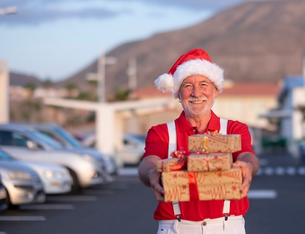 lächelnder älterer Mann, der für Weihnachten in einer Weihnachtsmütze und in Hosenträgern mit Geschenkboxen einkauft