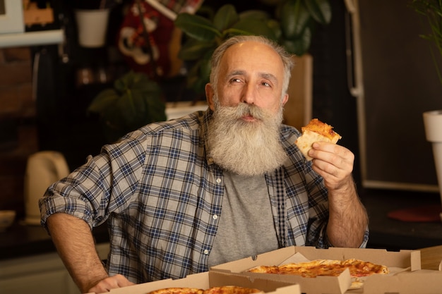 Lächelnder älterer Mann, der Pizzastück isst, das am Wohnzimmer sitzt. Bärtiger älterer Mann, der italienisches Essen isst.