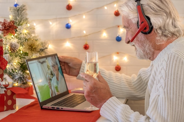 Lächelnder älterer Mann im Videoanruf mit Laptop, der Weihnachten mit einem Glas Sekt feiert. Frohes neues Jahr und frohe Weihnachten