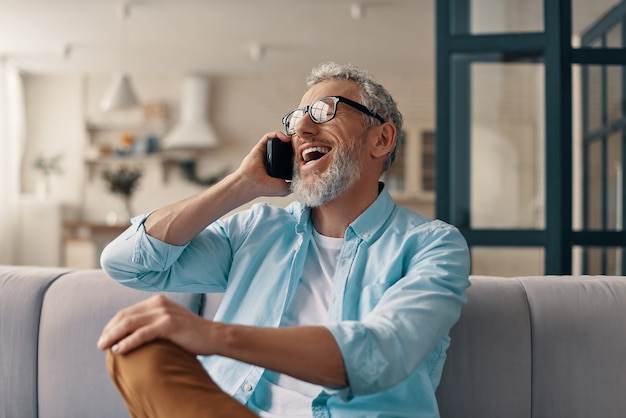 Lächelnder älterer Mann in Freizeitkleidung und Brille, der auf dem Smartphone spricht, während er zu Hause auf dem Sofa sitzt