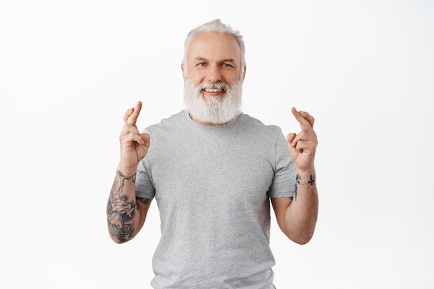 Lächelnder älterer Mann mit Tätowierungen, die Wünsche machen, optimistisch und hoffnungsvoll aussehen, auf gute Nachrichten warten, Traum wahr werden, in lässigem T-Shirt gegen weiße Wand stehen