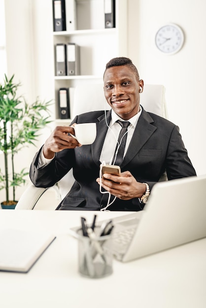 Lächelnder afrikanischer Geschäftsmann sitzt im Büro, trinkt Kaffee und hört Musik über Kopfhörer.