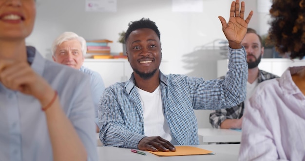 Lächelnder afrikanischer Student, der am Schreibtisch sitzt und im Klassenzimmer die Hand hebt