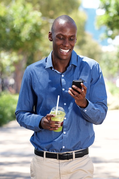 Lächelnder Afroamerikanermann, der draußen mit Mobiltelefon und Getränk geht