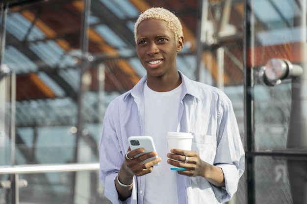 Lächelnder afroamerikanischer Hipster mit Handy, der eine Tasse Kaffee auf der Straße hält