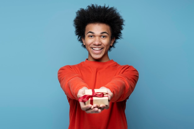 Lächelnder afroamerikanischer Mann mit Geschenkbox, der isoliert auf blauem Hintergrund in die Kamera blickt