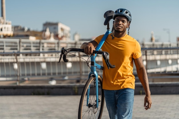 lächelnder afroamerikanischer Mann mit Helm hält sein Fahrrad und geht auf einer städtischen Straße