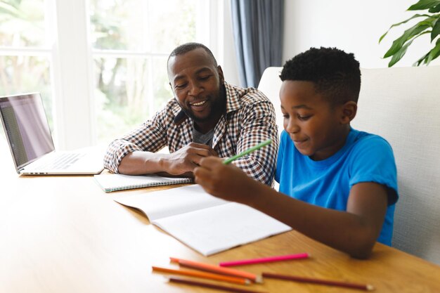 Lächelnder afroamerikanischer Vater und Sohn sitzen am Tisch im Esszimmer, arbeiten und machen Hausaufgaben