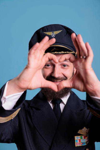 Lächelnder Airliner-Pilot, der herzförmiges Liebessymbol mit den Fingern zeigt, konzeptionelle Romantik-Geste. Freundlicher Flugzeugflieger in Uniform, der Zuneigungsgefühle ausdrückt, mittlerer Schuss