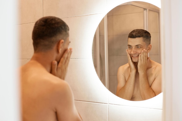 Lächelnder, angenehm aussehender, gutaussehender Mann mit nacktem Oberkörper, der vor dem Spiegel im Badezimmer steht und sein Gesicht berührt, nach der Rasur Lotion oder Balsam auf seine Wangen auftragen.