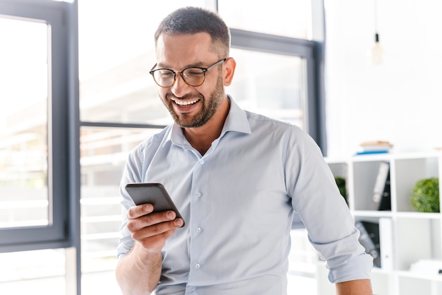 lächelnder Arbeitgebermann im weißen Hemd, der im Büroraum nahe großem Fenster steht und Smartphone für Arbeit verwendet