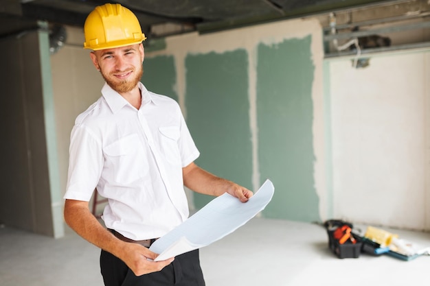 Lächelnder Architekt in weißem Hemd und gelbem Helm, der freudig auf den Plan der neuen Wohnung bei der Arbeit blickt