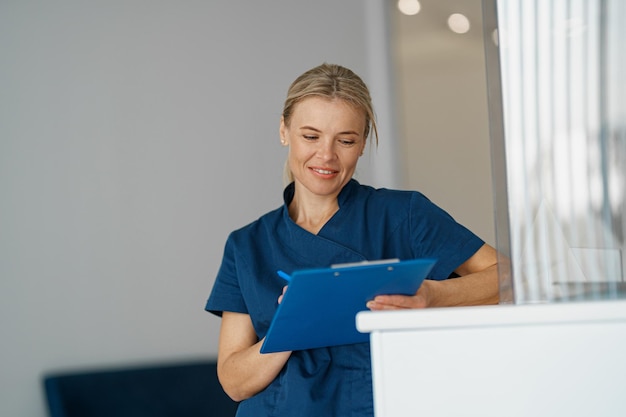 Lächelnder Arzt, der sich Notizen in der Zwischenablage macht, die in der Nähe der Rezeption in der Klinikhalle steht