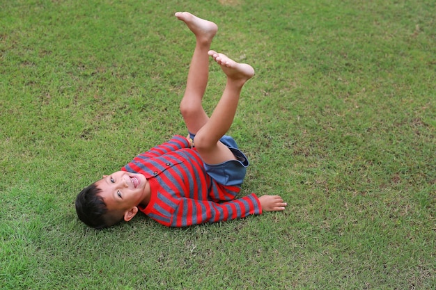 Foto lächelnder asiatischer kleiner junge, der auf grünem rasen liegt und seine beine hochhebt kid liegt auf gras mit suchender kamera
