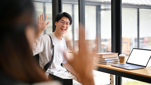 Lächelnder asiatischer männlicher Student winkt mit der Hand, um seinem Freund in einem Café Hallo zu sagen