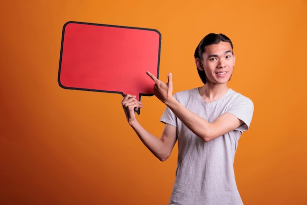 Lächelnder asiatischer Mann, der auf eine leere rote Sprachwolke mit Kopierraum zeigt, Werbespott oben. Fröhlicher Teenager mit Dialogblase, junger Mensch mit Nachrichtenrahmen, der in die Kamera schaut