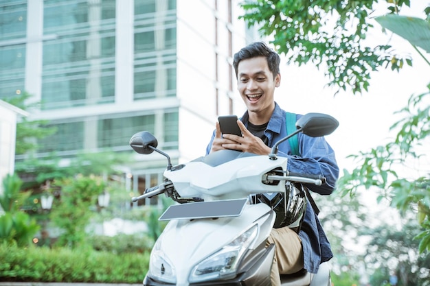 Lächelnder asiatischer Mann, der ein Mobiltelefon benutzt, während er ein Motorrad im Hintergrund der Stadtstraße fährt