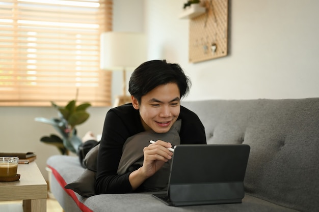 Lächelnder asiatischer Mann mit Computertablette auf dem Sofa zu Hause.